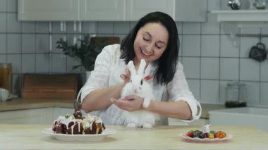 Beyaz elbiseli bir kadın mutfak masasında beyaz bir Paskalya tavşanını okşuyor. Küçük bir çocuk Paskalyayı kutluyor. Geleneksel Paskalya dini bayramı.