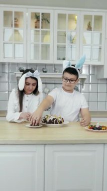 Kafasında tavşan kulağı olan bir kız ve erkek paskalya pastasını dekoratif yumurtalarla süslüyor. Paskalya pastası süslemesi. Mutlu Paskalyalar. Dikey video.