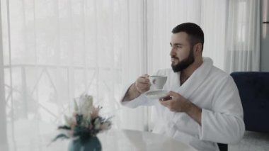 Beyaz cüppeli genç bir adam bir otel odasında bir masaya oturur, kahve içer ve pencereden dışarı bakar. Tatilde olan bir adam, sabah kahvesi sırasında güzel kış manzaralarına hayran olur..