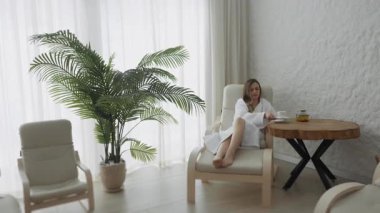 Beyaz cüppeli genç bir kadın spa salonunda oturuyor çay içiyor ve eğleniyor. Kız pencere kenarında oturur, sıcak bir şeyler içer ve pencereden dışarı bakar..