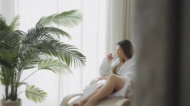 Beyaz cüppeli genç bir kadın spa salonunda oturuyor çay içiyor ve eğleniyor. Kız pencere kenarında oturur, sıcak bir şeyler içer ve pencereden dışarı bakar..