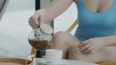 Mavi mayo giymiş bir kadın masaya oturur ve çaydanlıktan taze demlenmiş sıcak çayı fincana döker. Tatilde çay içen bir kız.