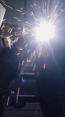  Koruyucu maskeli bir fabrikada çalışan bir işçi metal bir yapıyı kaynaklıyor. Metal yapılar üreten bir fabrika. Kaynak metalinden kıvılcımlar. Endüstriyel üretim. Dikey video.