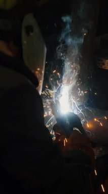  Koruyucu maskeli bir fabrikada çalışan bir işçi metal bir yapıyı kaynaklıyor. Metal yapılar üreten bir fabrika. Kaynak metalinden kıvılcımlar. Endüstriyel üretim. Dikey video.