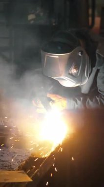 Bir fabrikada koruyucu maske takan bir işçi, elle tutulan bir plazma kesiciyle demir bir boruda delik açıyor. Metal yapılar ve ısıtma kazanları üretimi için endüstriyel fabrika. Dikey video.