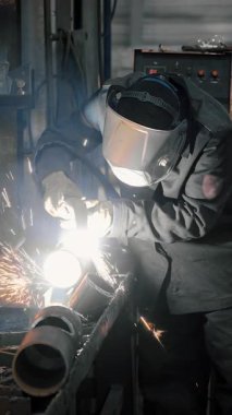 Bir fabrikada koruyucu maske takan bir işçi, elle tutulan bir plazma kesiciyle demir bir boruda delik açıyor. Metal yapılar ve ısıtma kazanları üretimi için endüstriyel fabrika. Dikey video.