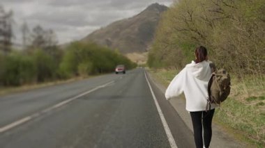 Dişi bir gezgin dağ yolunda otostop çekiyor ve yürüyor. Omuzlarında sırt çantası olan bir kız turist geçen arabalarla dağ yolunda yürüyor ve eliyle bir arabayı durdurmaya çalışıyor..