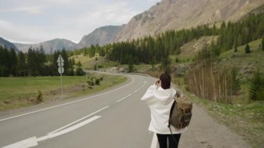 Sırtında sırt çantasıyla gezen genç bir kız dağ manzaraları arasında güzel bir yolda yürüyor ve manzaranın tadını çıkarıyor..
