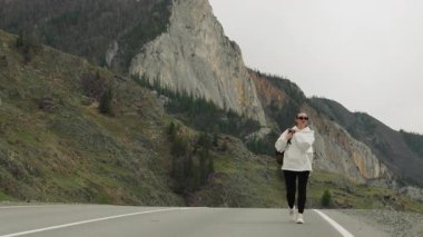 Sırtında sırt çantasıyla gezen genç bir kız dağ manzaraları arasında güzel bir yolda yürüyor ve manzaranın tadını çıkarıyor..