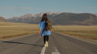 Dağlarda yol boyunca yürüyen bir kadının arka görüntüsü. Sırt çantasıyla yolda yürüyen bir kadın turist..