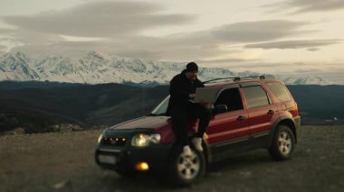 Dağın arka planında bir arabanın üzerinde otururken dizüstü bilgisayarda çalışan serbest çalışan bir erkeğin portresi. Elinde bilgisayarla arabanın kaputunda oturan bir erkek yolcunun silueti..