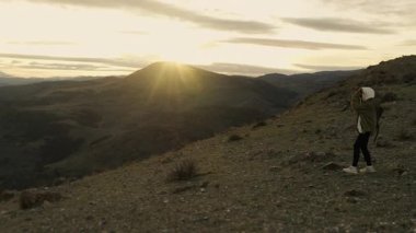 Sırt çantalı bir turist gün batımında dağların tepesindeki bir uçuruma yaklaşıyor ve dağların güzel panoramik manzarasının tadını çıkarıyor..
