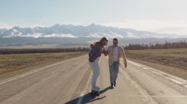 Bir çift genç turist, bir erkek ve bir kadın, dağ yolunda kaykay sürüyorlar. Adam kıza sörf tahtasına binmeyi öğretiyor. Dağlara aile gezisi..