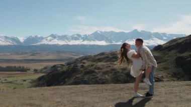 Birbirine aşık genç bir çift, bir erkek ve bir kadın, bir dağın tepesinde durup, güzel dağ manzarasına sarılıp öpüşüyorlar..