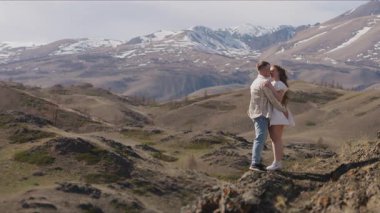 Birbirine aşık genç bir çift, bir erkek ve bir kadın, bir dağın tepesinde durup, güzel dağ manzarasına sarılıp öpüşüyorlar..