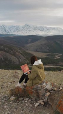 Elinde dizüstü bilgisayarı olan kırmızı ceketli bir kız gün batımında kar kaplı dağların zemininde bir taşın üzerinde oturuyor. Dağlarda uzaktan çalışan serbest çalışan genç bir kadın. Dikey video.