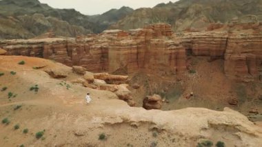 Beyaz elbiseli kadın turist, kırmızı kaya kanyonunda uçurumun kenarında koşuyor. İnanılmaz kırmızı kaya manzarasının tadını çıkarıyor. Panoramik drone manzarası..