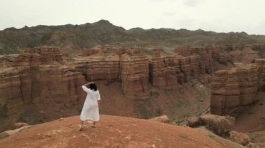 Beyaz elbiseli kadın turist, Red Rock Kanyonu 'nda uçurumun kenarında duruyor. İnanılmaz kırmızı kaya manzarasının tadını çıkarıyor. Panoramik drone manzarası..