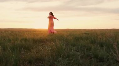 Pembe elbiseli ve uzun saçlı mutlu genç bir kadın gün batımında sahada ellerini kaldırarak mutlu anların tadını çıkarıyor..