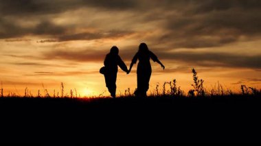 Gün batımında el ele tutuşarak tarlada koşan mutlu anne ve kız siluetleri. Doğada aile tatili.