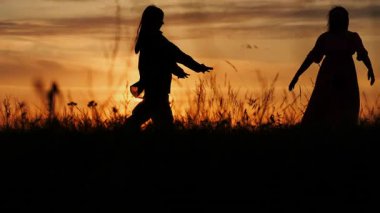 Gün batımında bir tarlada el ele tutuşan mutlu anne ve kız siluetleri. Doğada aile tatili.