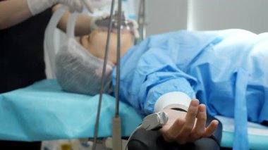 Bir kadın hasta, bir klinikte ameliyat masasında yatıyor. Parmağında nabız monitörü var. Doktor, solunum maskesi takıyor..
