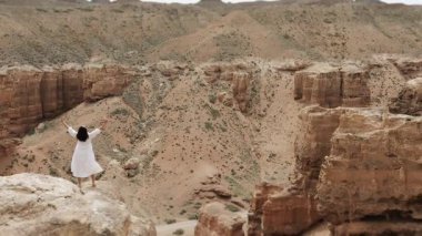 Kırmızı kaya kanyonunda uçurumun kenarına yürüyen beyaz elbiseli kadın turist ellerini kaldırıyor ve inanılmaz kırmızı kaya manzarasının tadını çıkarıyor..