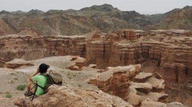 Şapkalı ve sırtında bir sırt çantası olan küçük bir turist bir dağın tepesinde oturur ve kanyonun güzel manzarasına hayran kalır..