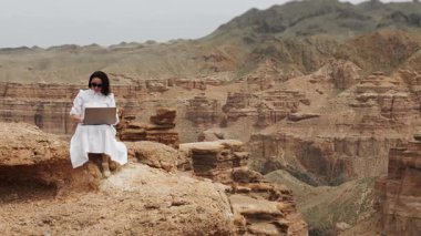 Beyaz elbiseli bir kadın turist, uçurumun kenarında dizüstü bilgisayarıyla oturuyor ve uzaktan kumandayla yazı yazıyor..