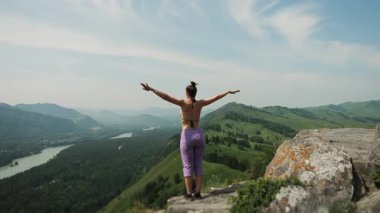 Güzel, genç, sıska bir kız yoga pozisyonunda duruyor. Ellerini dağın tepesine kaldırıyor. Yeşil yaz dağlarının güzel manzarasının tadını çıkarıyor..