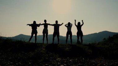 Gün batımında bir tarlada duran beş genç turistin siluetleri elleri havada dağlarda tatillerinin tadını çıkarıyorlar..