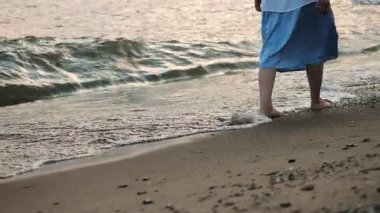 Denizden kumsala doğru yürüyen narin bacaklı bir kadının yakın çekimi. Deniz dalgaları bacaklarına çarpıyor..