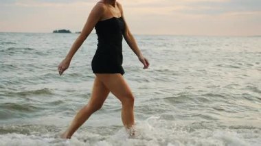 Mayolu genç bir kız tropik bir sahilde yalınayak yürüyor, deniz dalgaları kızlara çarpıyor. Denizde tatil yapan bir kadın turist..