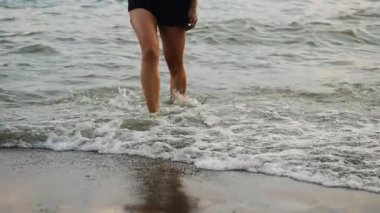 Denizden kumsala doğru yürüyen narin bacaklı bir kadının yakın çekimi. Deniz dalgaları bacaklarına çarpıyor..