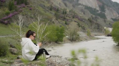 Genç, güzel bir turist kız dağlarda bir kayaya oturur ve ısınmak için çelik termostan sıcak bir içecek döker. Kız dağlarda yürüyüş yaparken dinleniyor..