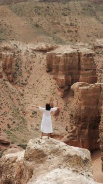 Kırmızı kaya kanyonundaki uçurumun kenarına doğru yürüyen beyaz elbiseli kadın turist ellerini kaldırıyor ve inanılmaz kırmızı kaya manzarasının tadını çıkarıyor. Dikey video.