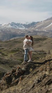 Birbirine aşık genç bir çift, bir erkek ve bir kadın, dağın tepesinde durup, güzel dağ manzarasına sarılıp öpüşüyorlar. Dikey video.