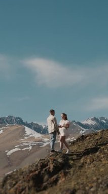 Birbirine aşık genç bir çift, bir erkek ve bir kadın, dağın tepesinde durup, güzel dağ manzarasına sarılıp öpüşüyorlar. Dikey video.