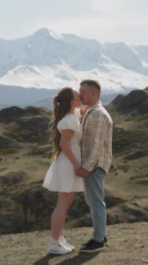 Birbirine aşık genç bir çift, bir erkek ve bir kadın, dağın tepesinde durup, güzel dağ manzarasına sarılıp öpüşüyorlar. Dikey video.