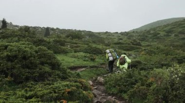 İki turist, bir erkek ve bir kadın, sırt çantalı ve yürüyüş direkli yağmurluklu, bir dağın tepesine dik bir yamaçtan tırmanıyorlar..