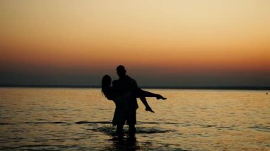 Gün batımında, denizden kıyıya bir kızı kollarında taşıyan bir adamın silueti. Denizde tatilde olan sevgi dolu bir çift.
