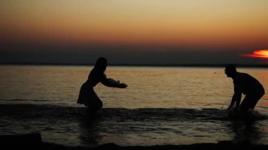 Gün batımında denizde dururlarken birbirlerine su sıçratan bir erkek ve kadının siluetleri. Denizde sevgi dolu bir çiftin aile tatili.