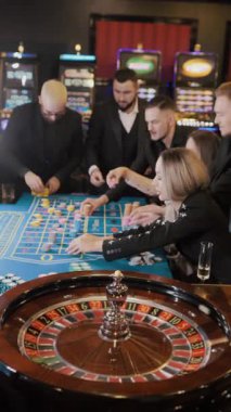 Bir grup genç kadın ve erkek kumarhanede rulete oynuyorlar. Kumarhanede kumar oynamak, oyuncuların oyun sahasına çip koyması. Dikey video.