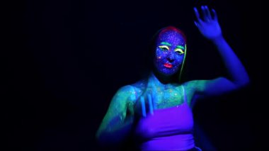 Karanlık arka planda floresan renklerle boyanmış genç bir kızın portresi. Parlak makyajlı bir kadın kameraya poz verir..