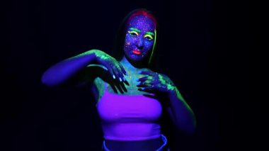 Karanlık arka planda floresan renklerle boyanmış genç bir kızın portresi. Parlak makyajlı bir kadın kameraya poz verir..