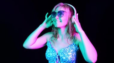 Güneş gözlüklü ve kulaklıklı mutlu bir kızın portresi neon ışıkta müzik dinliyor. Neon ışıkta dans eden kız.