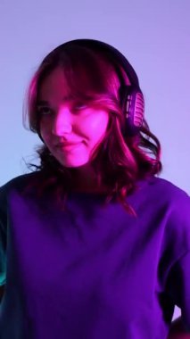Güneş gözlüklü ve kulaklıklı mutlu bir kızın portresi neon ışıkta müzik dinliyor. Neon ışıkta dans eden kız. Dikey video.