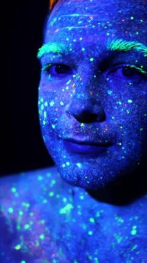 Kameraya bakan floresan boyalarla boyanmış bir adamın yakın çekimi. Parlak makyajlı bir yüz. Dikey video.