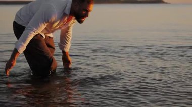 Gün batımında elleri su sıçratan, tişörtlü vahşi bir adamın silueti. Bir erkek turist denize su sıçratıyor..