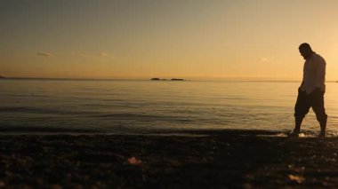 Gün batımında deniz kıyısında yürüyen, gömlekli vahşi bir adamın silueti. Bir adam kumsalda yürür ve gün batımının tadını çıkarır..